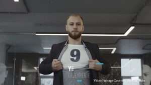 Bearded man in a blazer pulls open his shirt to reveal a large '9' on a white tee, in an office setting.